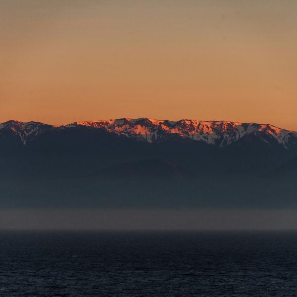 A breathtaking view of a snow-covered mountain range during sunset with a calm ocean in the foreground.