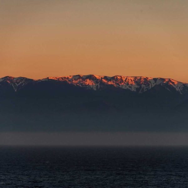 A breathtaking view of a snow-covered mountain range during sunset with a calm ocean in the foreground.